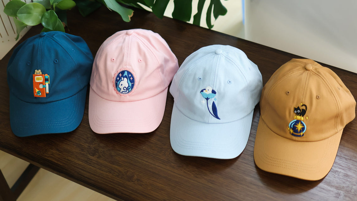 Four baseball caps in blue, pink, white, and yellow on a wooden surface with plants in the background.