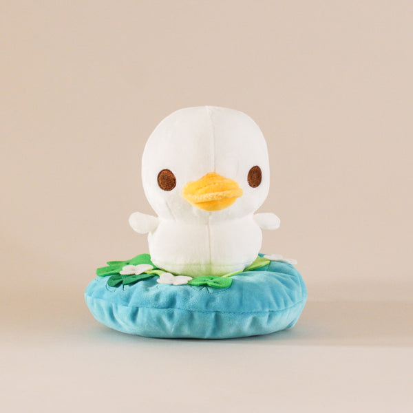 A lost Duckling Plushie. White colored duck swimming on top of small pond with lily pads and small flowers
